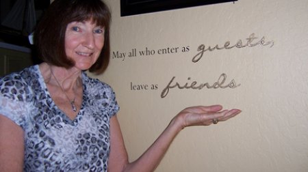 Photo of Anita Bevan next to plaque saying "May all who enter as guests leave as friends."