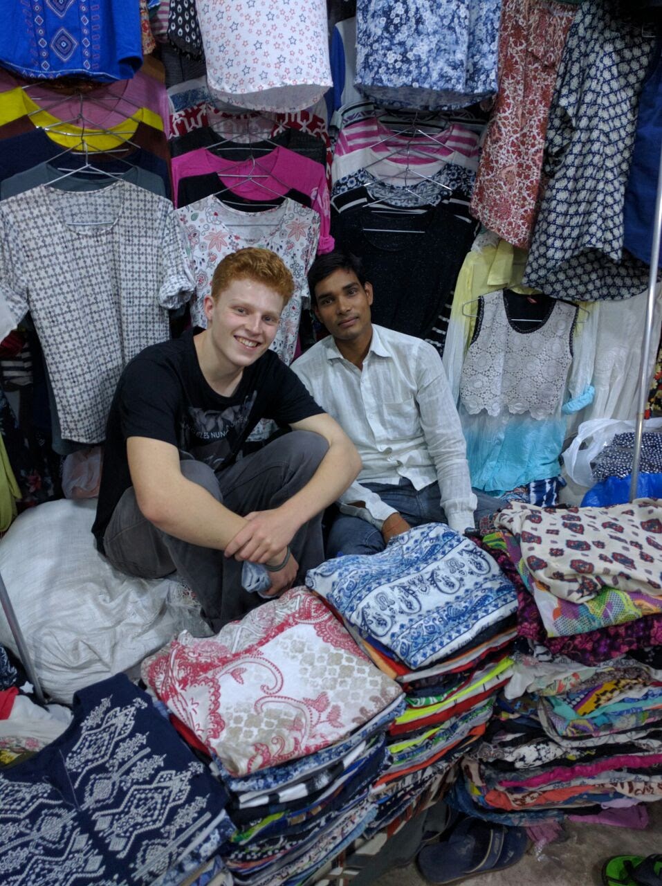 Young guy sits next to a man in the middle of fabric piles and clothes hanging in a store
