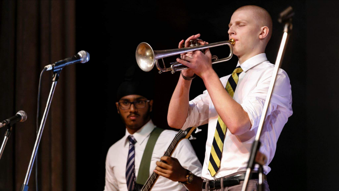 Young man plays trumpet on a stage in front of a mic with another young man in the background on a guitar