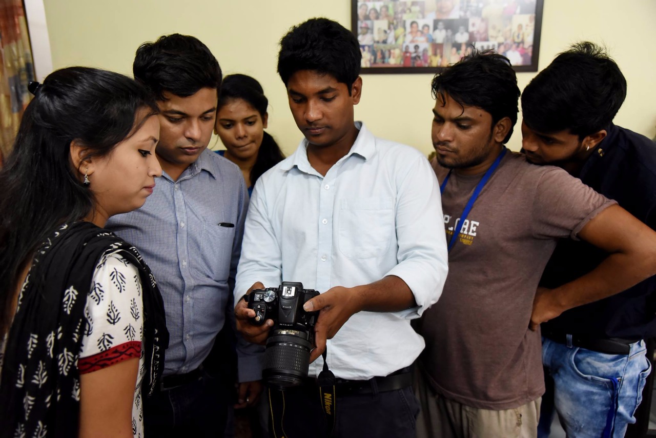 Young man holding a fancy camera surrounded by a group of young adults observing his actions with the camera