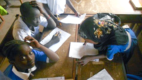 Three young children working at a desk, one is looking up at camera