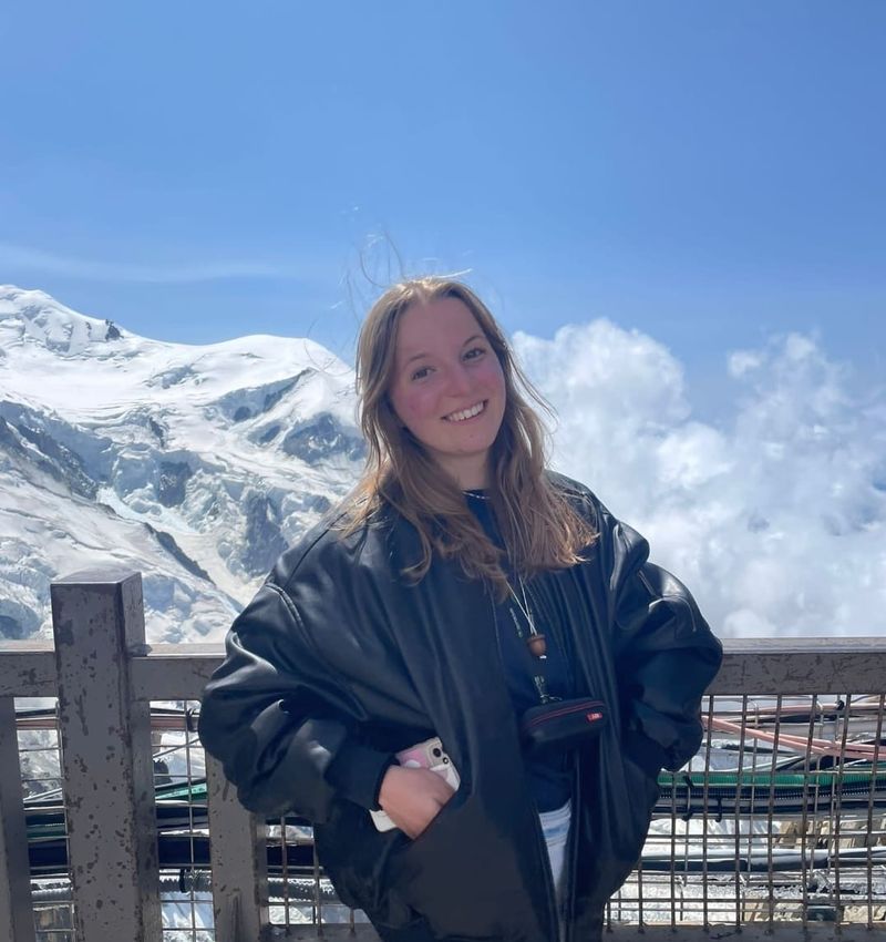 A photo of Julia posing near a fence, with a mountain backdrop behind her