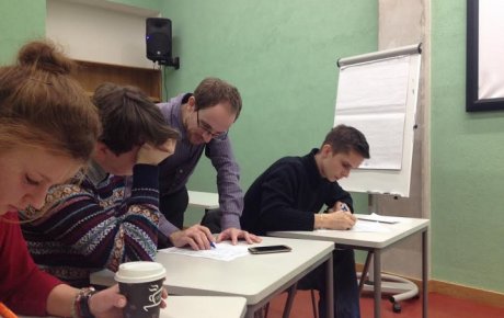 Three students at desks in a classroom with male teacher leaning over one desk