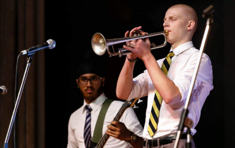 Young man plays trumpet on a stage in front of a mic with another young man in the background on a guitar