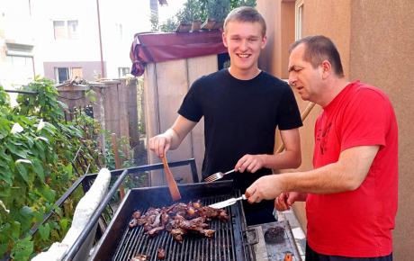 Teenage boy smiling and helping flip meat on a grill with an older man