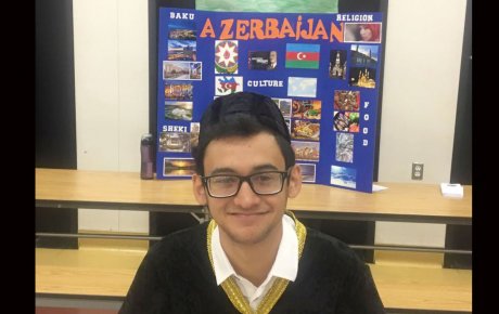 Young man wearing glasses and baseball hat to the back, sits smiling in front of display on Azerbaijan
