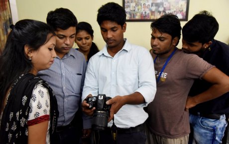 Young man holding a fancy camera surrounded by a group of young adults observing his actions with the camera