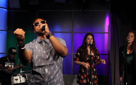 Man holding mic wearing sunglasses and holding his fist out as he sings on stage with background singers behind him