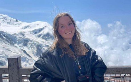 A photo of Julia posing near a fence, with a mountain backdrop behind her