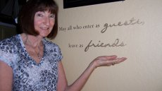 Photo of Anita Bevan next to plaque saying "May all who enter as guests leave as friends."