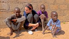 Young woman sits on dirt ground against brick wall with 3 young boys that are not wearing shoes