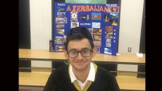 Young man wearing glasses and baseball hat to the back, sits smiling in front of display on Azerbaijan