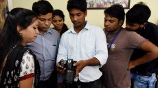 Young man holding a fancy camera surrounded by a group of young adults observing his actions with the camera