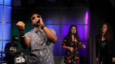 Man holding mic wearing sunglasses and holding his fist out as he sings on stage with background singers behind him