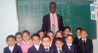 Male teacher wearing blue and red (USA colors) tie stands smiling with a group of young children in front of chalkboard